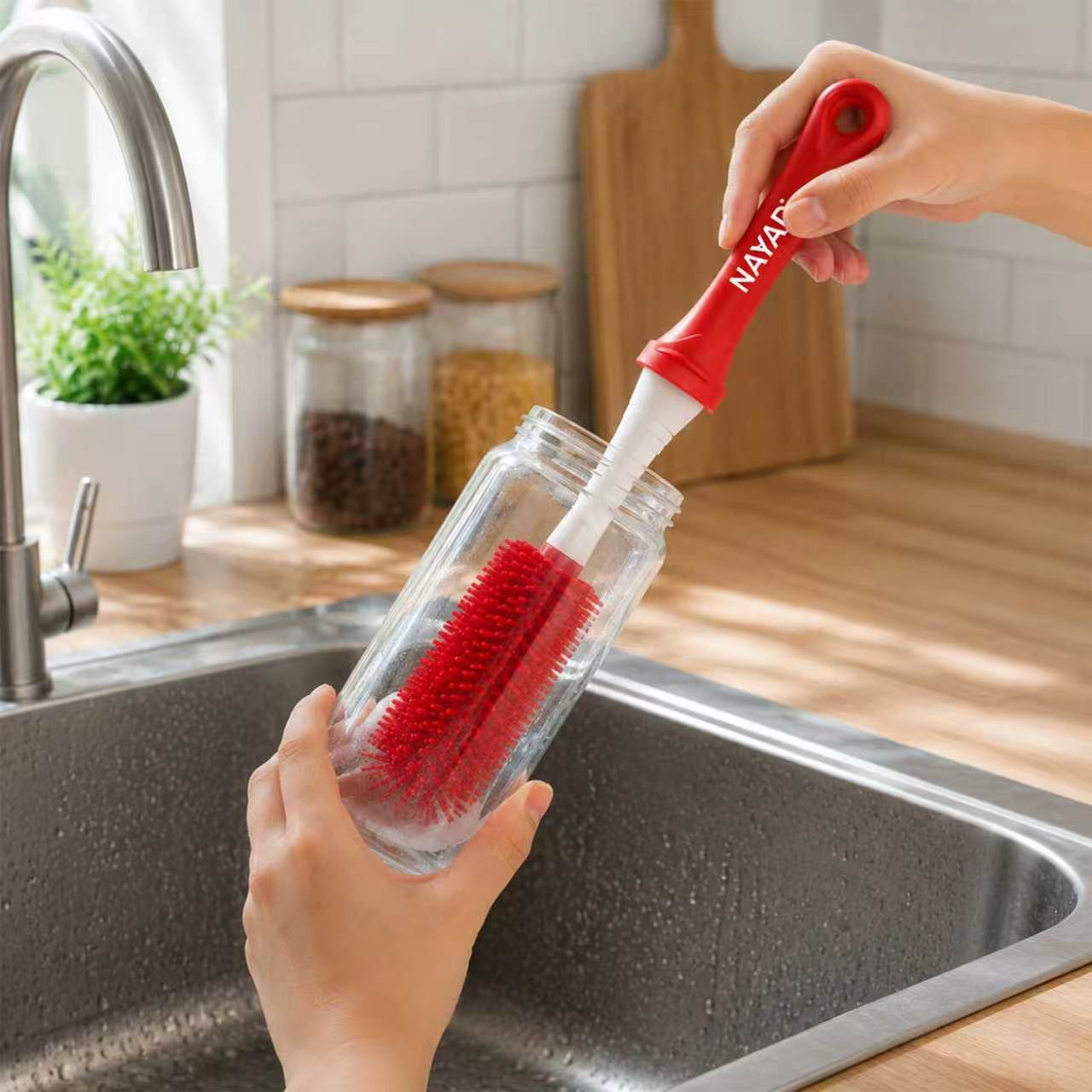 A red and white cleaning brush with a clear plastic handle is being used to clean a clear glass bottle in a kitchen sink. The brush has a red handle and red bristles, with the brand name 'NAYAD' printed on the white section of the handle.