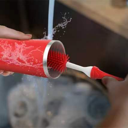 A close-up of a red silicone container being rinsed under running water with a red-and-white cleaning brush inside. A hand holds the container and another hand holds the brush, with a sink and faucet visible in the background.