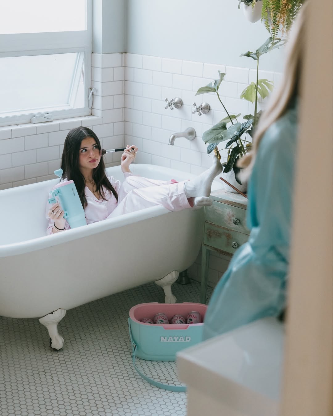 Woman in a bathtub with a blue tumbler, surrounded by plants and a towel rack.