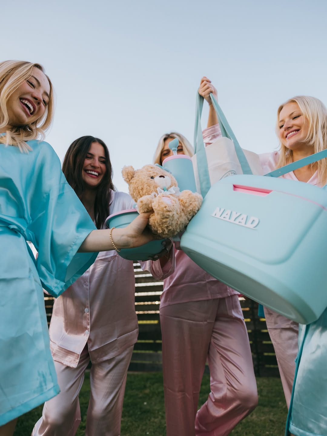 Four women in matching outfits holding a teddy bear and a blue cooler set with 'NAYAD' branding.