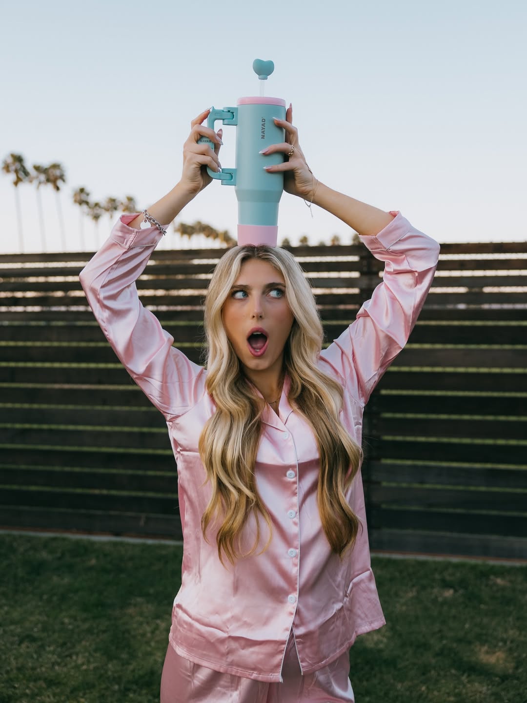 Person in pink pajamas holding a colorful mug above their head outdoors.