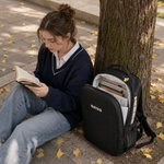 A woman sits on a stone bench reading a book next to a tree, with a black NAYAD backpack placed on the ground beside her. The backpack is open, showing a light gray interior with books inside, and the scene is outdoors with fallen yellow leaves on the ground.