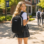 A smiling young woman on a college campus wearing a black NAYAD backpack, a white shirt, and a plaid skirt, with other students and campus buildings blurred in the background.