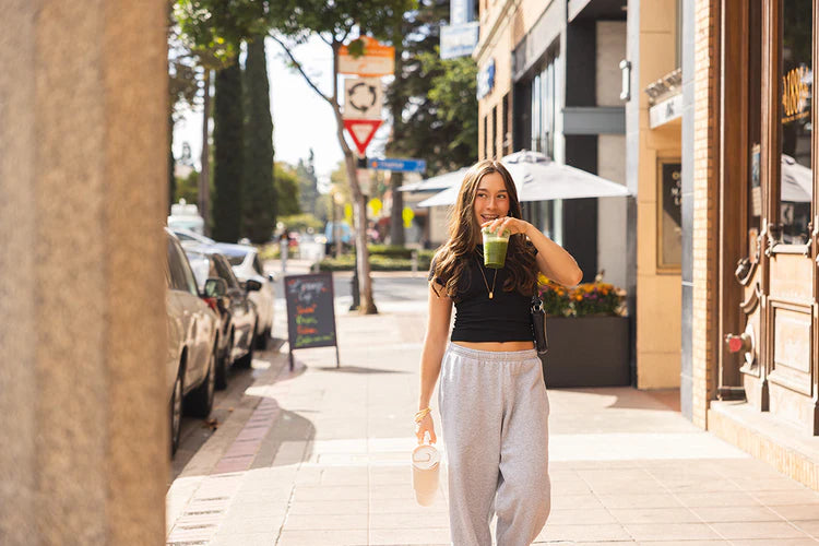 Woman walking down a city street holding a drink, with storefronts and pedestrians in the background.