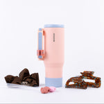 A tall insulated tumbler in pastel pink and baby blue accent with a handle, styled on a white background with a brown scrunchie, extra straws, and hair clips.