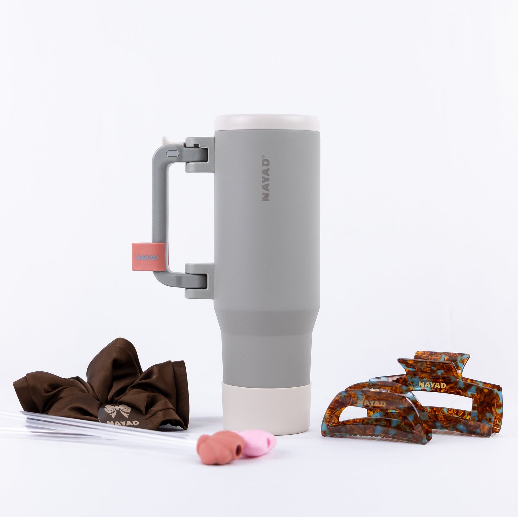A gray and white insulated travel tumbler with a handle, shown on a white background with styling accessories including a brown hair scrunchie, extra straws, and hair clips.