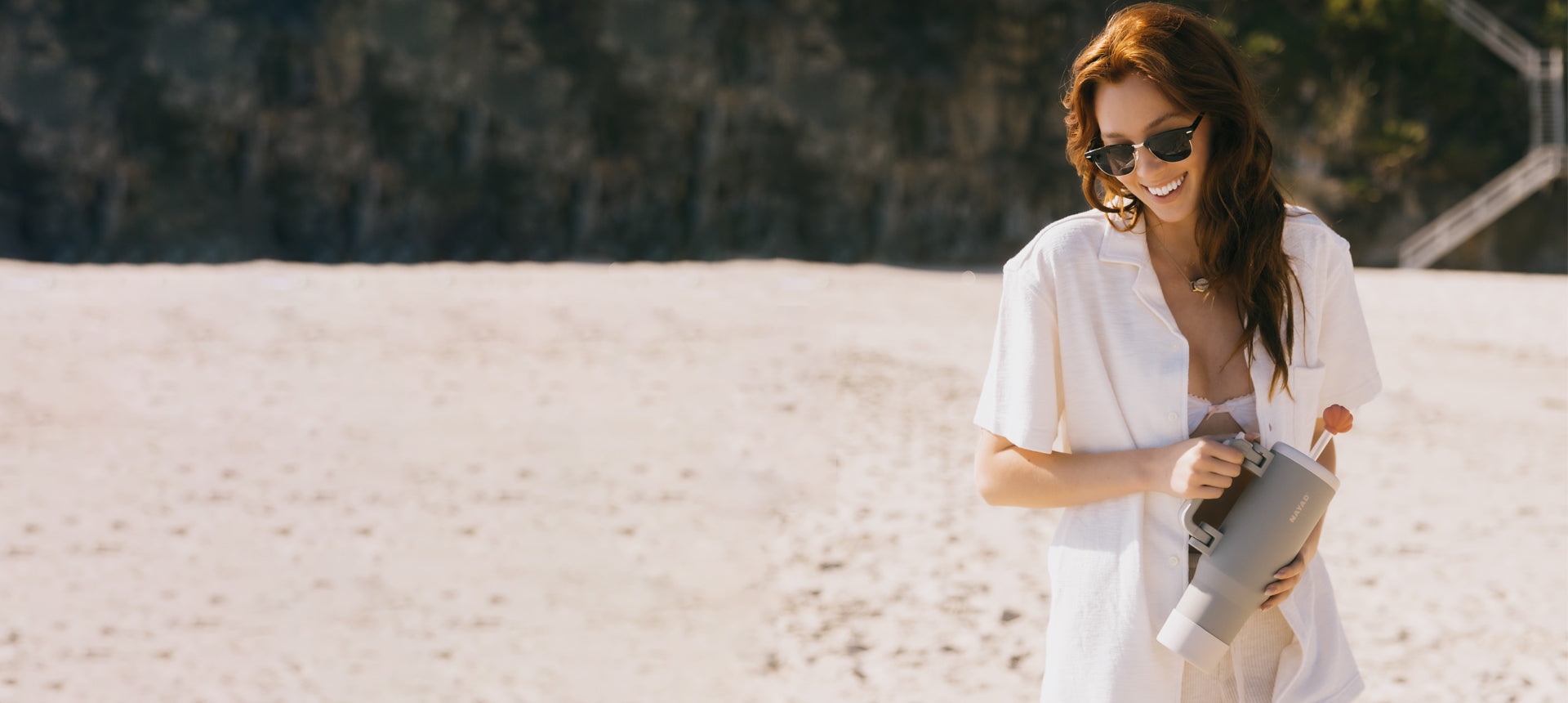 Woman in a white dress holding a gray tumbler on a sandy beach.